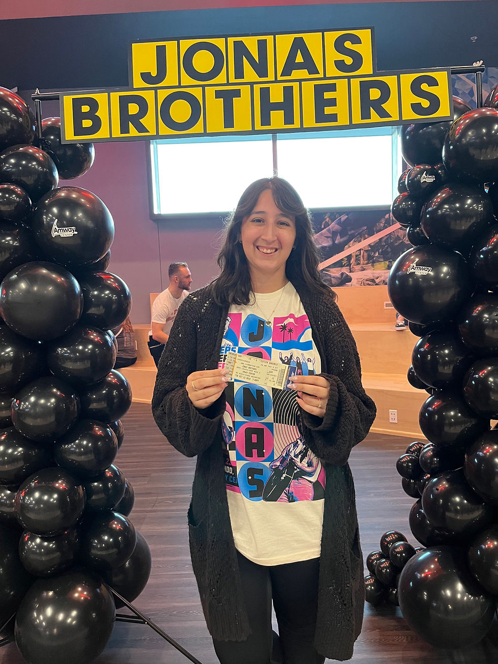 Person smiling, holding a ticket under a "Jonas Brothers" sign with black balloon decorations. Casual setting with a colorful shirt.