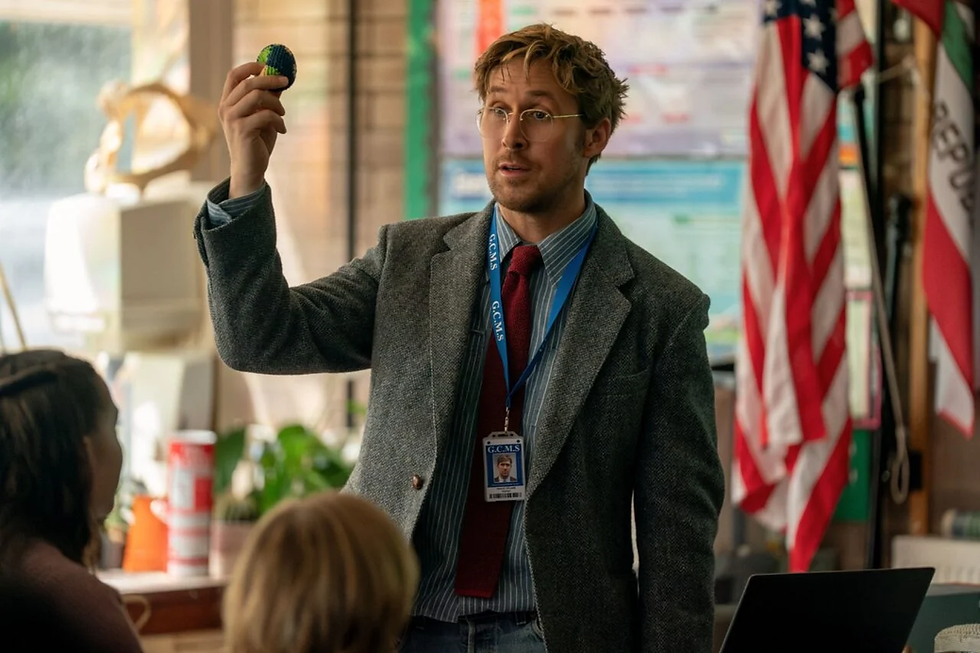 Teacher in blazer holds up a small knitted ball in a classroom, wearing a G.C.M.S. badge, with U.S. flags behind him.