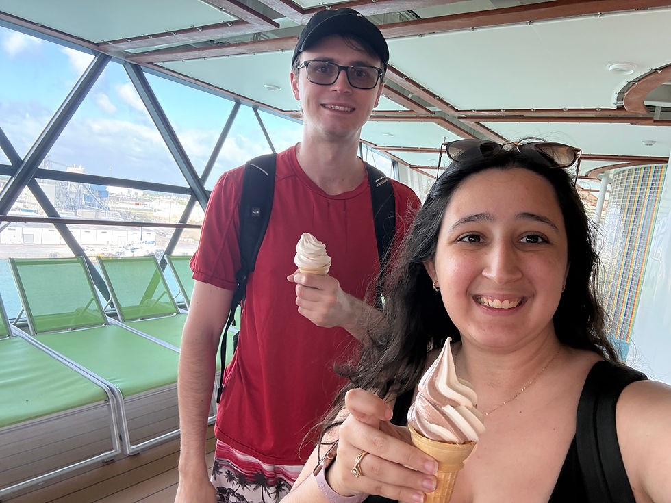 Two people smiling with ice cream cones on a ship deck. Green lounge chairs, glass roof, and ocean view in the background. Casual, happy mood.