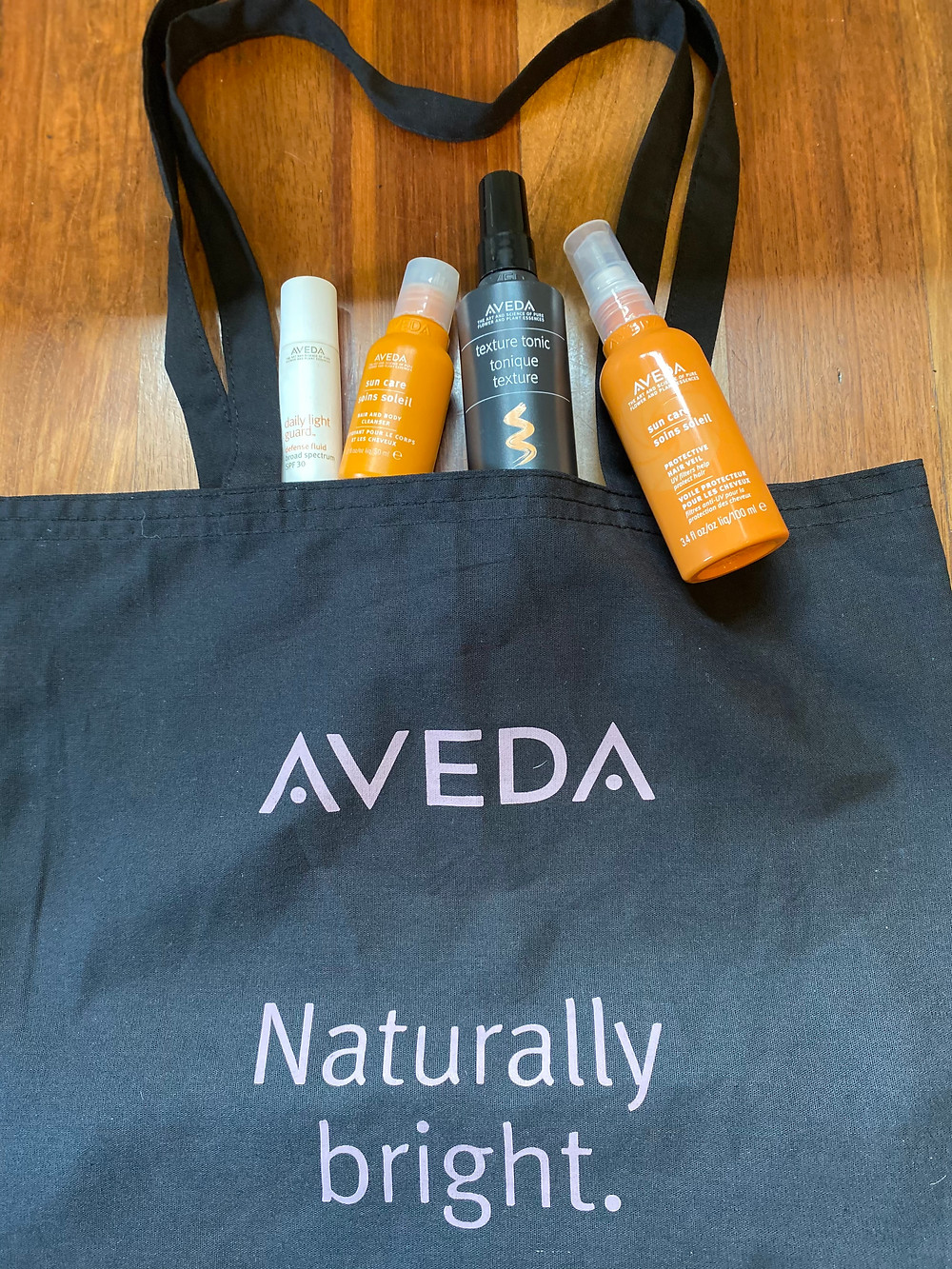 Aveda Naturally Bright tote bag with Aveda products Daily Light Guard, Sun Care Hair Veil, Texture Tonic, and travel size of Sun Care Hair and Body Wash
