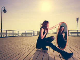 Woman sitting on a boardwalk looking into a standing mirror with the sun shining behind her.
