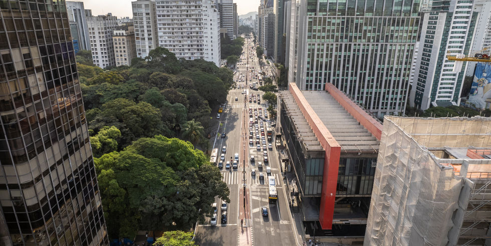Vista aérea da Avenida Paulista durante o dia, mostrando edifícios altos e áreas verdes ao longo da avenida.