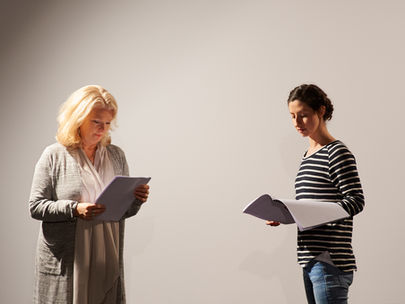 Two women standing in a white room. They have scripts in their hands that they are reading