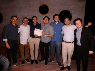 Seven smiling men pose indoors, one holding a certificate. They wear casual shirts and pants. The background features a gray wall with circular mirrors.