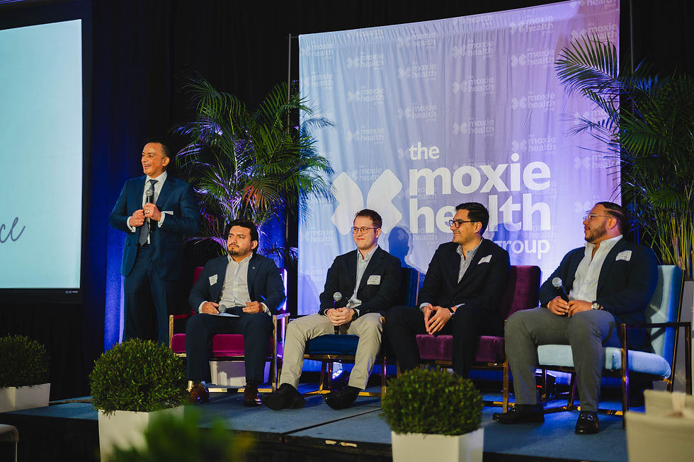 Panelists on stage at the Moxie Health Group event; one man speaks while four sit with microphones amid palms.