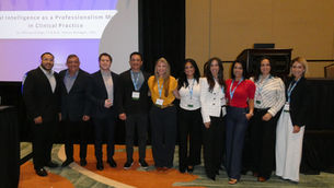 Conference group poses before AI in Clinical Practice slide at a hotel podium; mixed formal attire, smiling.