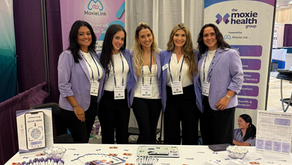 Five women in lavender blazers smile at a booth with Moxie Health banners. Promotional items are displayed on the table.