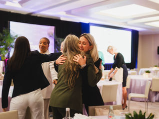 Women hugging at a conference in a warmly lit room. Others chat and organize materials at round tables. Mood is friendly and welcoming.