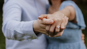 Close-up of a couple holding hands, showing a diamond engagement ring. The background is blurred, and both wear light blue clothing, evoking romance.