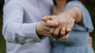 Close-up of a couple holding hands, showing a diamond engagement ring. The background is blurred, and both wear light blue clothing, evoking romance.