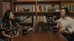 Two people with headphones converse in a cozy podcast studio. Bookshelves filled with colorful books are in the background.