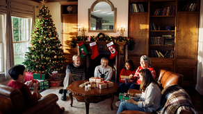 Family opening gifts by a decorated Christmas tree and fireplace with stockings. Cozy room with wood shelves, festive mood, and warm lighting.