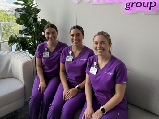 Three women in purple scrubs smile while sitting on a couch inside an office with a leafy plant. A neon sign is partially visible above.