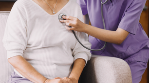 Healthcare worker in purple scrubs uses stethoscope on seated woman in white top. Indoor setting with calm mood, beige armchair.