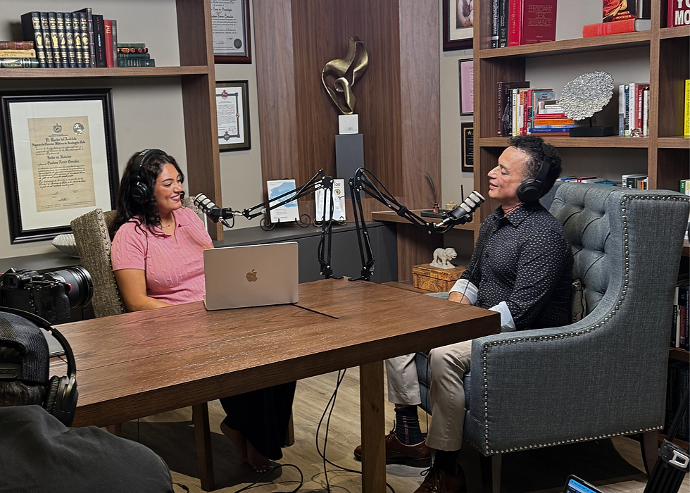 Two people recording a podcast at a wooden table with microphones. Bookshelves and certificates decorate the background. Both appear engaged.