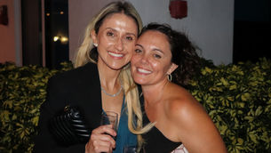 Two women smiling at the camera, holding drinks, in front of green foliage. The mood is cheerful and celebratory.