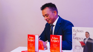 Man in a blue suit signs copies of "Echoes of Legacy" at a table. Red books are stacked. Mood is professional and focused.