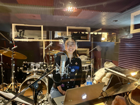 I am sitting in a recording booth filled with instruments and a mic with a black cardigan on that has fishes on it. T