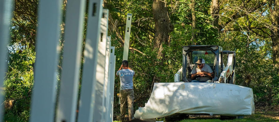 Two workers in a wooded area install white fence posts. One operates machinery, the other positions a post. Sunlight filters through trees.