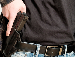 Close-up of a person wearing jeans and a black shirt, holstering a handgun at the waist. They are adjusting the gun in a black belt.