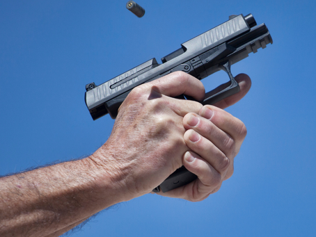 Hands firing a black pistol against a clear blue sky, with an ejected bullet casing mid-air above the gun