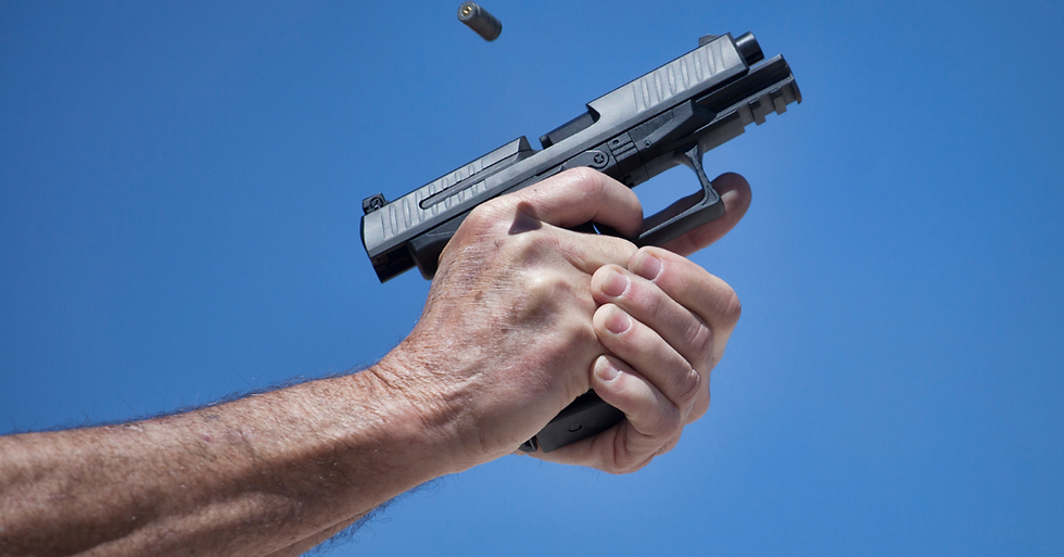 Hands firing a black pistol against a clear blue sky, with an ejected bullet casing mid-air above the gun.
