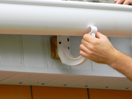 Hands install a white gutter to a roof edge using a bracket. Background shows white siding. Overcast lighting creates a neutral mood.