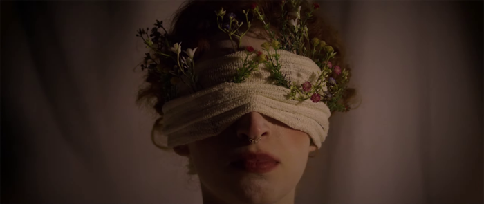 Frame do clipe. Rosto de uma mulher branca com uma atadura enrolada no rosto na altura dos olhos pela qual saem flores.