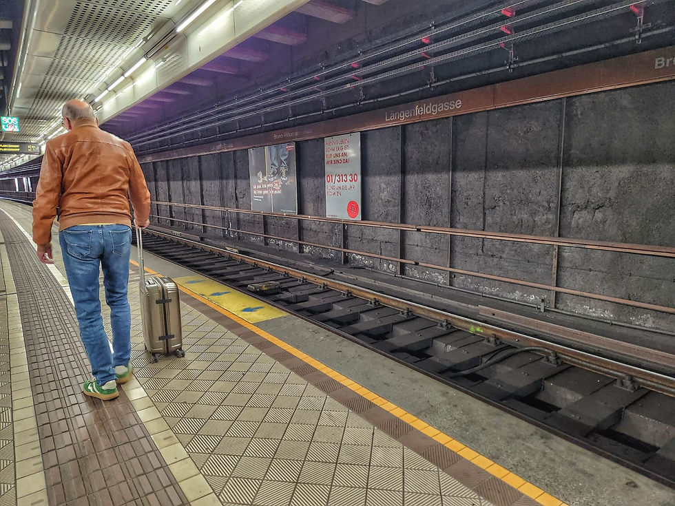 Man in a brown jacket walks on a subway platform with a suitcase. Signs read "Längenfeldgasse." Mood is calm, setting is urban.