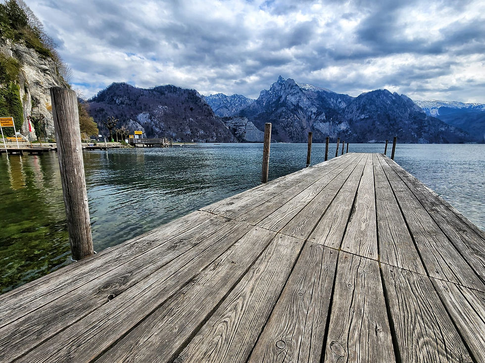 Wooden dock extending into a calm lake, surrounded by mountains. Cloudy sky and greenish water create a tranquil atmosphere. Signs visible.