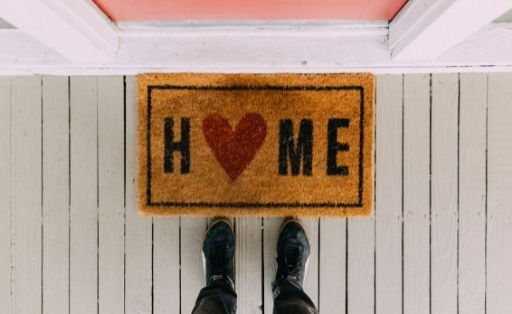 A door mat with the word home on it, by a front door, with someone standing near beside the mat, with only their shoes and ankles in the image