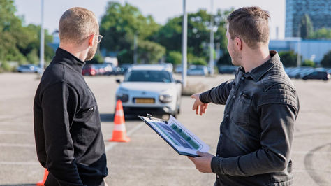 Dva muškarca razgovaraju na parkiralištu, dok jedan od njih pokazuje prema automobilu i čunju u pozadini.