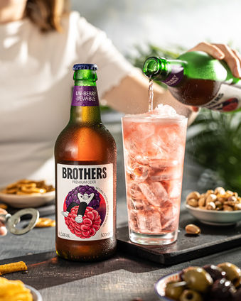  Brothers cider bottle in a glass with a pink tint to the cider. Tapas is on the table surrounding the products and a woman is pouring the product into a glass. Shot to look like they are outside, but shot in a creative Bristol food and drink photography studio