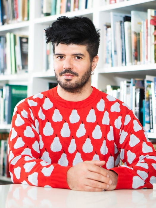 a man wearing a red jumper is sitting at a table with his hands clasped together. He is sitting in front of a bookshelf and looking into the camera