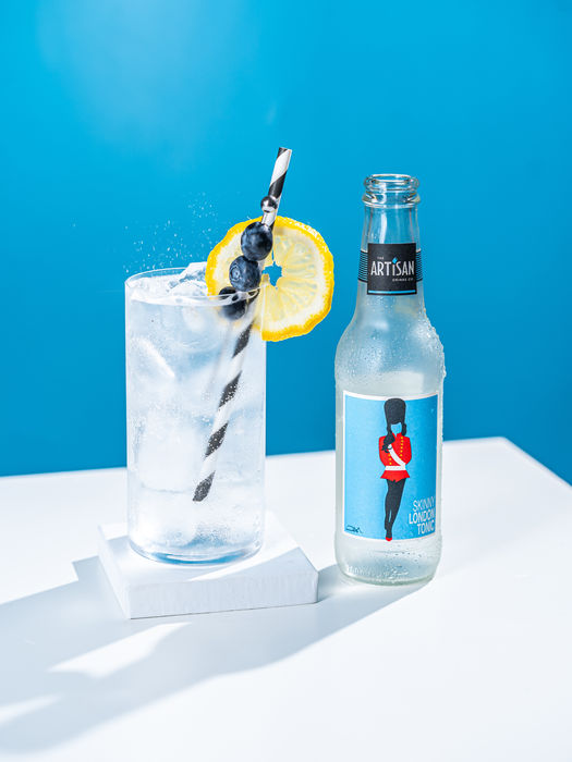 a bottle of tonic water and a full glass of gina and tonic with a slice of lemon and a straw have been placed on a table with a blue background. shot at bristol studio Future Proof product and spirit photography