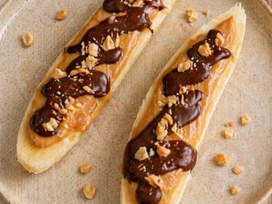 Bananes fraîches garnies de beurre de cacahuète artisanal La Pistache de Terre, nappées de chocolat noir fondu et parsemées de cacahuètes grillées, présentées sur une assiette beige.