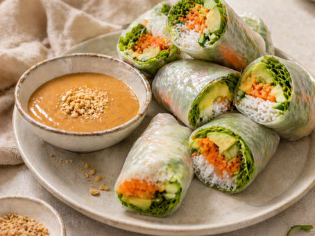 Rouleaux de printemps frais garnis d’avocat, carotte, concombre et vermicelles de riz, servis avec une sauce onctueuse au beurre de cacahuète. Présentation élégante sur assiette en céramique dans des tons beige clair, style culinaire raffiné et minimaliste, idéal pour illustrer une recette saine et gourmande.