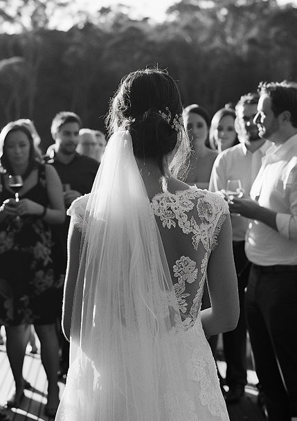 A bride in a lace gown seen from behind as she approaches a crowd of guests, emotion captured in wedding photos