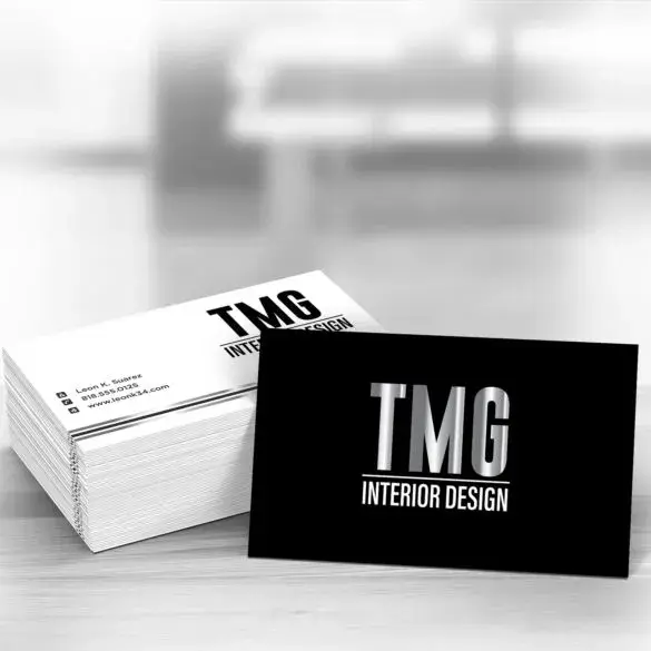 Stacked white business cards with black text and a silver foil logo 'TMG INTERIOR DESIGN' against a light background.