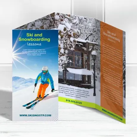 A quad-fold brochure for "Ski and Snowboarding lessons" stands partially open. The front cover shows a skier on a sunny mountain slope, while the inside panels display a snowy cabin scene and an orange section with text.