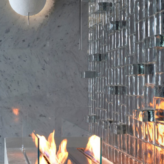 ▊在這處新店住宅中,起居室主牆採用了透明實心玻璃磚打造隔間牆,不僅作為空間的分界,也成為整體設計的視覺亮點。選用圓角水波光2010款式,利用膠築立砌與膠築躺砌雙向工法,層層堆疊、錯落排列,構築出凹凸造型排列的立體牆面,讓每一束光線在磚面上反射出不同的表情。
此案最大特色,在於整面牆皆由透明實心玻璃磚拼砌而成,透光卻不透明,提供隱私之餘,也保有光線穿透的通透感。透過森森團隊的施工專業技術,克服了磚體重量與排列精度的挑戰,使玻璃磚牆體在細節中展現工藝美感,穩固又安全。
凹凸造型排列打破了單一平面的呆板視覺,使整體牆面不僅止於隔間功能,更具有如藝術裝置般的美學存在。隨著日光與燈光變化,牆體光影層次活潑豐富,彷彿為靜謐空間注入流動感,讓每一次停留與注視,都能感受到不同的光影氛圍。
這是一面兼具結構、光感與藝術性的實心玻璃磚牆,是住宅空間裡不可忽視的靈魂主角。