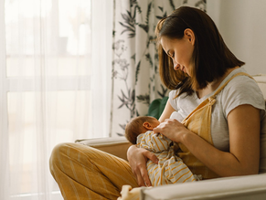 A woman in yellow overalls breastfeeds a baby in a cozy room. Soft light filters through sheer curtains, creating a warm, peaceful mood.