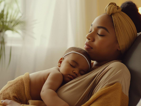 A woman with a tan headwrap holds a sleeping baby on her chest in a cozy room with soft lighting and a leafy plant in the background.