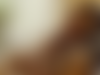 A woman with a tan headwrap holds a sleeping baby on her chest in a cozy room with soft lighting and a leafy plant in the background.