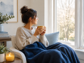Woman in a cozy sweater holds a yellow mug, gazing out a bright window. Blue blanket and pillow, book stack, candle, and plant nearby. Calm mood.