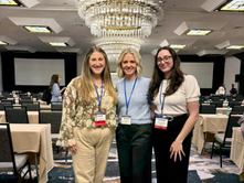 Three women wearing lanyards pose smiling in a conference ballroom with chandeliers, tables, and chairs in the background.