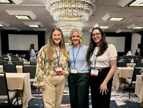 Three women wearing lanyards pose smiling in a conference ballroom with chandeliers, tables, and chairs in the background.