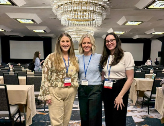 Three women wearing lanyards pose smiling in a conference ballroom with chandeliers, tables, and chairs in the background.