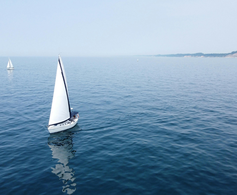 A white sailboat glides on calm blue sea under a clear sky, with distant cliffs and another sailboat visible in the background.