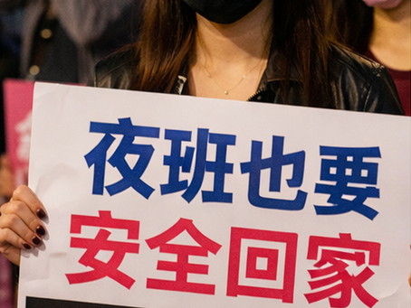 夜色中,定點茶站女員工戴著口罩、手持大型標語牌,站在群眾與媒體鏡頭前。標語以醒目字體強調「夜班也要安全回家」與「爭取夜間交通補助」,將個人通勤焦慮轉化為公共議題,象徵夜間勞動者以集體行動要求制度回應。這張影像呈現的是一種「把風險說出口」的瞬間:在夜晚工作的勞動者不再用沉默承擔危險,而是選擇以請願、連署、站上街頭的方式,要求城市把夜間通勤視為公共安全與勞動保障的一部分。背景中若隱若現的攝影者與同伴,也暗示倡議行動的媒體化與集體性——當訴求被看見,制度才可能被迫回應。